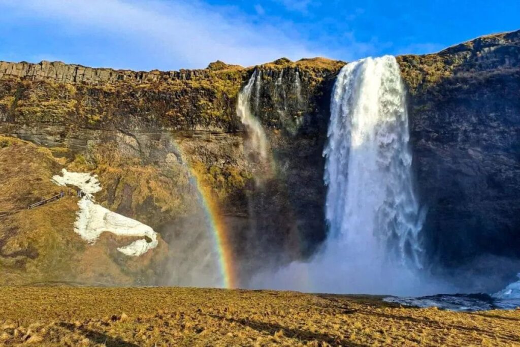 A powerful waterfall cascades over a rocky cliff into a misty basin below. The main column of water plunges straight down from the top of the cliff, while a smaller stream flows beside it along the rock face. The surrounding cliffs are covered in mossy green and brown vegetation typical of volcanic landscapes. Mist from the falling water rises into the air and catches the sunlight, forming a bright rainbow that arches upward from the base of the waterfall. The ground in the foreground appears rocky and textured, with patches of earth and sparse vegetation. On the left side of the cliff, a narrow walking path with a railing can be seen, allowing visitors to approach the waterfall safely. The scene captures the dramatic beauty of a large natural waterfall surrounded by rugged terrain and vibrant natural colors.