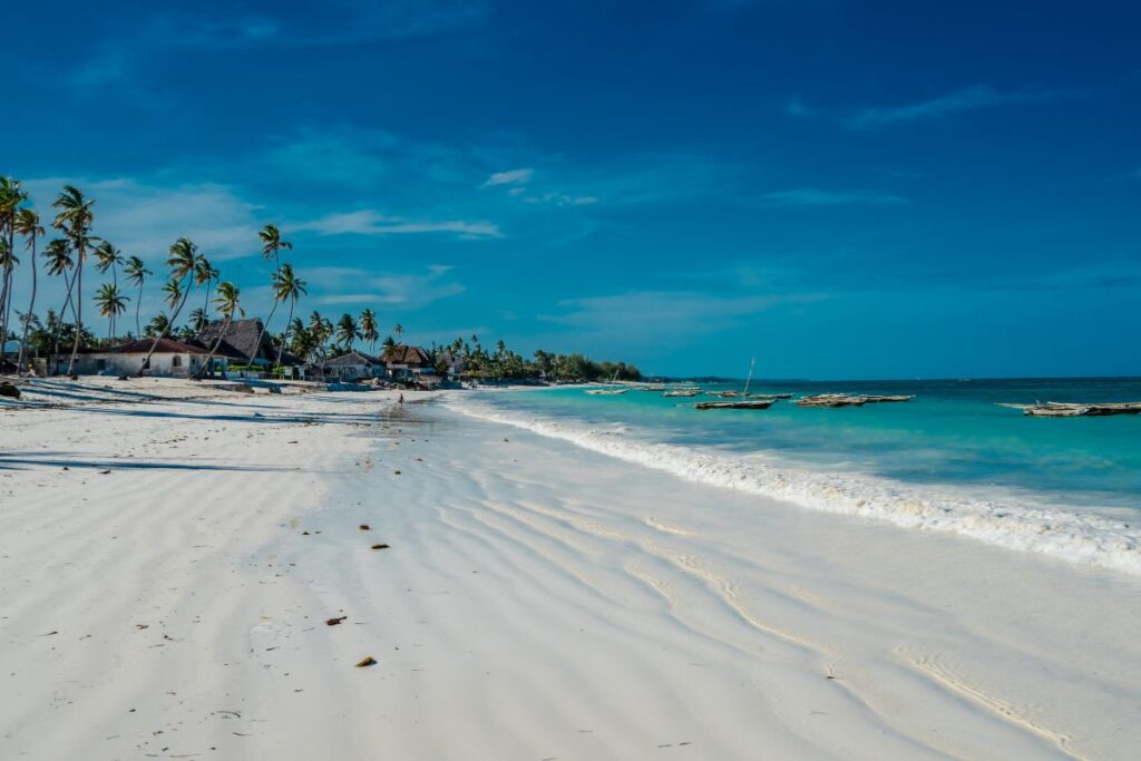 A long stretch of bright white sand beach extends along the shoreline beside clear turquoise ocean water. Gentle waves roll onto the sand, leaving smooth patterns where the water has receded. Several traditional wooden boats float close to the shore in the shallow water. Along the beach, tall palm trees lean slightly toward the sea, their green fronds swaying above small beachfront huts and buildings set back from the sand. The sky above is a vibrant blue with very few clouds, creating a warm and sunny tropical atmosphere. The coastline curves gently into the distance, where more palm trees and buildings line the shore. The image captures a peaceful tropical beach setting with calm water, soft sand, and coastal vegetation typical of many island destinations.