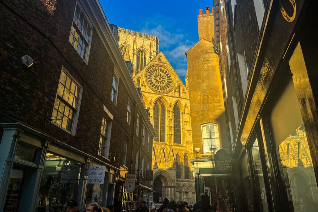 York Minster Street View A narrow historic street leads toward the towering facade of York Minster cathedral glowing in warm golden light. The Gothic cathedral rises dramatically at the end of the street, with tall pointed arches and an intricate circular rose window dominating the upper facade. The sunlight illuminates the stone, creating a strong contrast against the deep blue evening sky. On both sides of the street, small historic buildings house shops and cafes with hanging signs and display windows. Several pedestrians walk along the street, some pausing to look toward the cathedral. The tight alleyway frames the massive structure, emphasizing its height and detailed architecture. The scene captures a striking moment when the low sun casts golden light across the historic cathedral.
