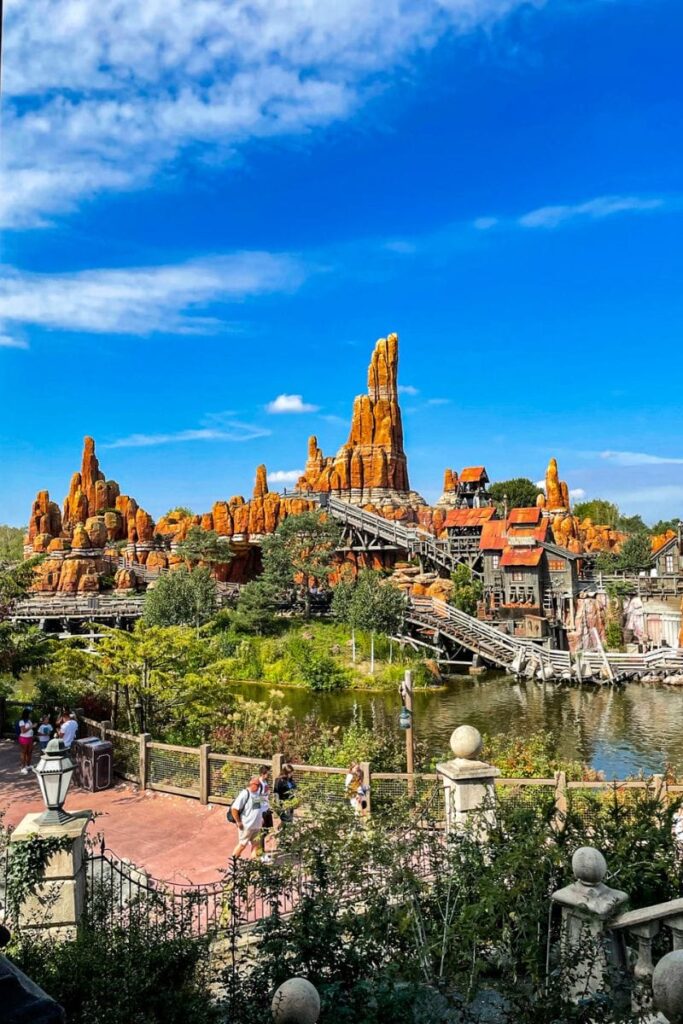 Big-Thunder-Mountain The image shows a large theme park attraction designed to resemble a rugged desert landscape with tall, jagged orange rock formations rising dramatically against a bright blue sky. Wooden roller coaster tracks weave through the rocks, supported by beams and trestles that add to the rustic, mining-town theme. In the foreground, a calm body of water reflects the surrounding scenery, bordered by green shrubs and trees. A pedestrian pathway runs along the lower edge of the image, where several visitors walk past decorative railings and lampposts. Small wooden buildings with weathered textures and slanted roofs sit near the base of the rocks, enhancing the illusion of an old frontier town. The scene is vibrant and detailed, combining naturalistic landscaping with themed architecture.