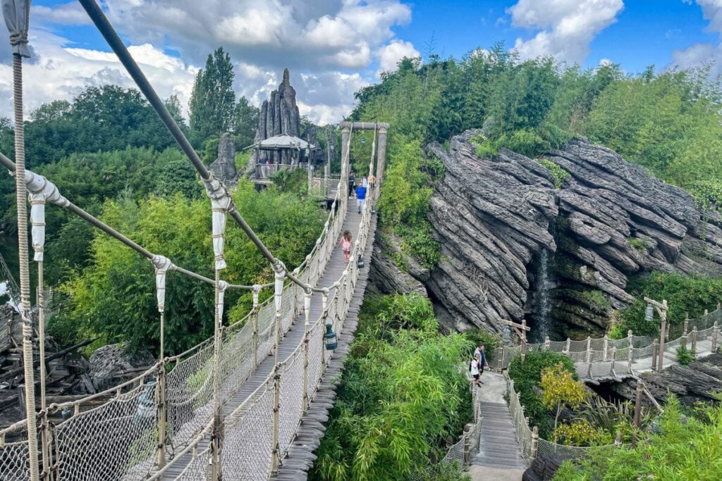 This image features a rope suspension bridge stretching diagonally across the frame, suspended above a lush, green landscape. The bridge is made of wooden planks with rope handrails and netted sides, creating a slightly adventurous, jungle-like feel. Several people are walking across it, spaced out along its length. Below and around the bridge, dense vegetation including bamboo and leafy trees fills the scene. To the right, a large rock formation with layered textures rises beside a narrow pathway that curves beneath the bridge. In the distance, a small lookout structure or platform is partially visible among the greenery. The sky above is partly cloudy, casting soft, diffused light across the scene. The overall atmosphere feels immersive and exploratory.