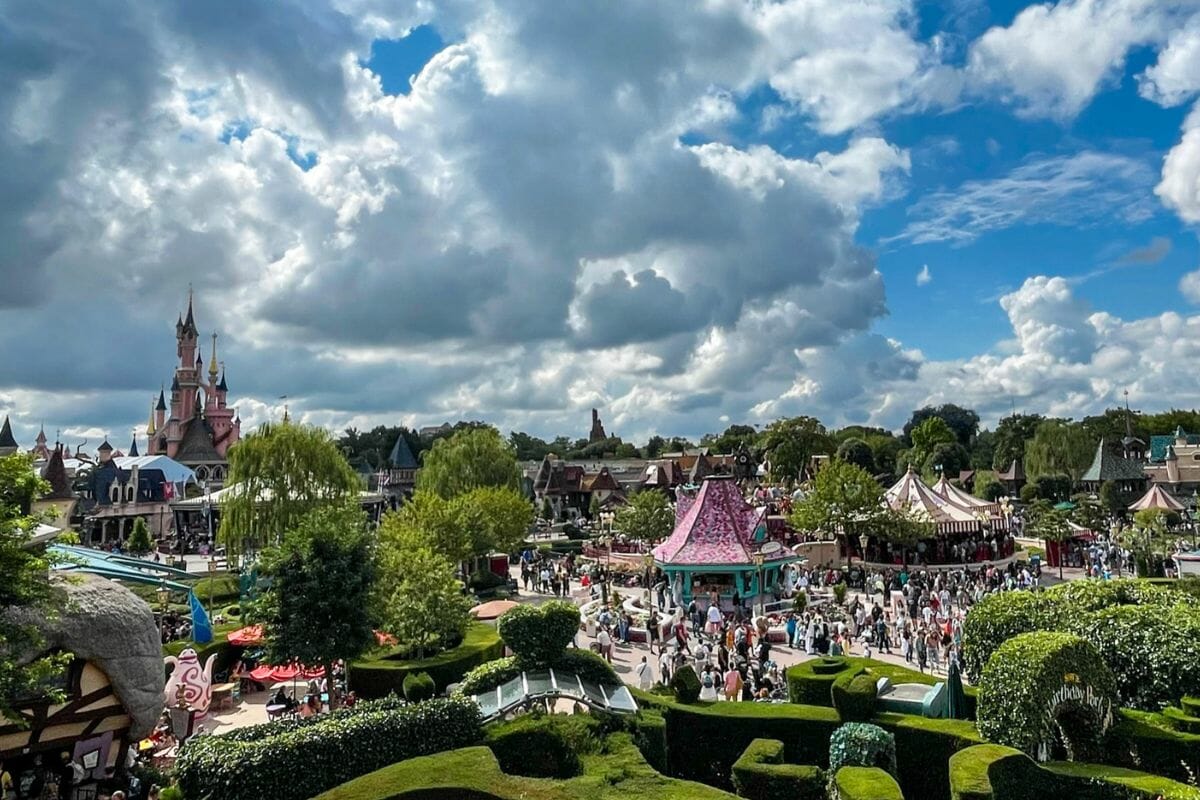 The image presents a wide, elevated view over a bustling theme park filled with visitors. In the distance to the left, a pink fairytale-style castle with tall spires stands as a focal point. The foreground features neatly trimmed green hedges and landscaped gardens with curved pathways. Colourful buildings with whimsical designs and pointed roofs surround a central plaza area, where large crowds of people gather and move between attractions. The sky is filled with dramatic, fluffy clouds against a blue backdrop, adding depth to the scene. Trees and decorative elements are scattered throughout, creating a lively and vibrant atmosphere. The image captures the scale and energy of the park, with a mix of architecture, greenery, and crowds.