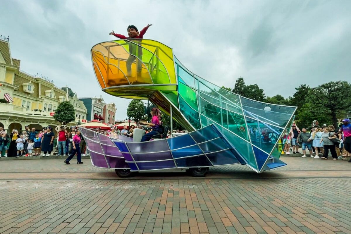 The image shows a colourful parade float moving along a paved street, surrounded by spectators on both sides. The float has a modern, geometric design with curved panels in bright gradient colours, including green, yellow, blue, and purple. A performer stands on an elevated section of the float with arms raised, appearing joyful and energetic. Another performer is seated below, possibly controlling the float. The crowd nearby includes families and children watching and taking photos. Buildings with classic theme park architecture line the background, featuring pastel colours and decorative details. The sky is overcast, creating softer lighting that contrasts with the vivid colours of the float. The scene conveys movement, celebration, and a festive parade atmosphere.