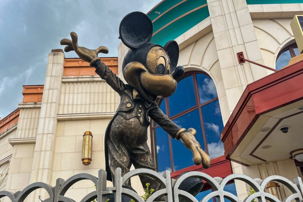 This image shows a bronze statue of Mickey Mouse standing behind a decorative metal fence. Mickey is posed with one arm extended outward and the other slightly raised, as if greeting visitors. The statue has a slightly weathered metallic finish that reflects light subtly. Behind the statue, a large building with cream-coloured walls, tall windows, and architectural detailing provides a structured backdrop. A section of the building features teal and red accents, adding contrast to the neutral tones. The sky above is cloudy and grey, giving the scene a softer, muted light. The composition places the statue prominently in the foreground, making it the focal point, while the building adds context and scale.