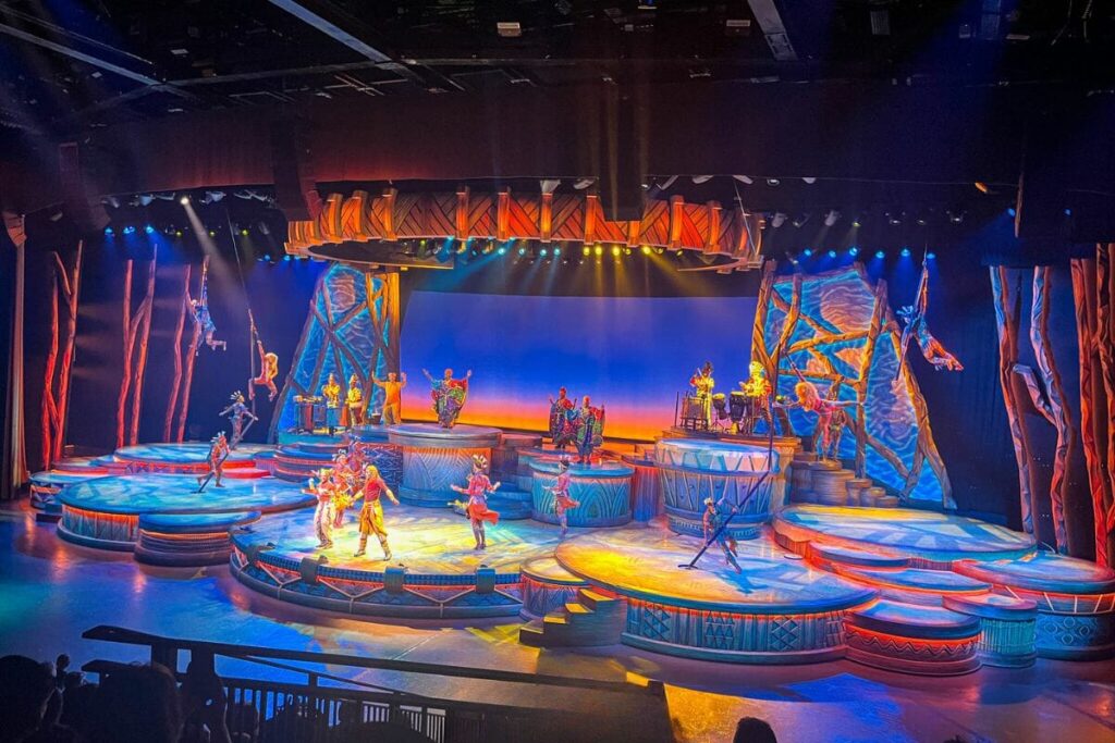 Lion-King-Show-at-Disneyland-Paris The image captures a large indoor theatre stage filled with performers in vibrant costumes. The stage is designed with multiple circular platforms at different heights, creating depth and dimension. Bright, colourful lighting in shades of blue, orange, yellow, and purple illuminates the scene, highlighting dancers and musicians positioned across the stage. Some performers appear mid-dance, while others stand on elevated platforms or play instruments. The backdrop includes stylised set pieces resembling natural or tribal elements, with bold shapes and textures. Overhead stage lights cast beams across the performance area, enhancing the dramatic effect. The audience seating is partially visible in the foreground, framing the performance. The overall scene feels energetic, theatrical, and visually rich.
