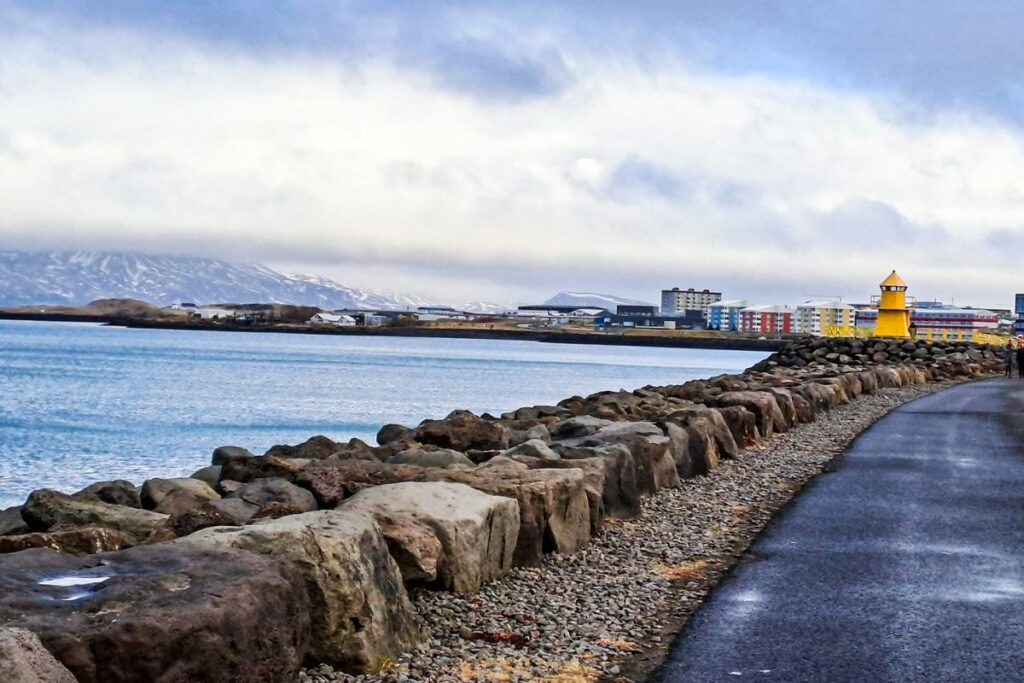 Reykjavik, Iceland-2 The image shows a coastal walking path running alongside a rocky shoreline, with large, uneven boulders forming a barrier between the paved walkway and the calm water. The water is a muted blue-grey, reflecting the overcast sky above. In the distance, low-rise buildings line the waterfront, including a prominent small yellow lighthouse with a bright orange top, standing near the edge of the shore. Beyond the buildings, mountains rise under a thick layer of clouds, with patches of snow visible on the slopes. The path curves gently to the right, bordered by gravel and rocks, creating a textured foreground. The scene feels quiet and peaceful, with soft natural light diffused by the cloudy sky. The combination of water, mountains, and coastal infrastructure creates a serene, expansive landscape.