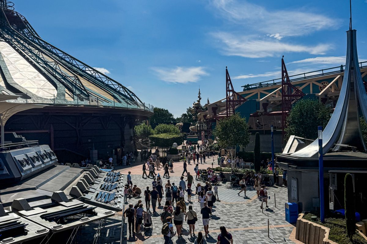 The image shows a busy futuristic-themed area inside a theme park on a bright sunny day. Large metallic structures dominate both sides of the walkway, featuring sleek silver surfaces, angular panels, and curved architectural elements that create a science-fiction atmosphere. In the centre of the image, wide paved walkways are filled with visitors walking in different directions, including families with pushchairs and groups of friends. Trees and landscaped greenery soften the futuristic surroundings, while additional attractions and structures are visible further in the distance. The sky is vivid blue with thin, wispy clouds stretched across it, and strong sunlight casts long shadows on the ground. The overall scene feels energetic and immersive, combining modern architectural design with the lively atmosphere of a crowded theme park.