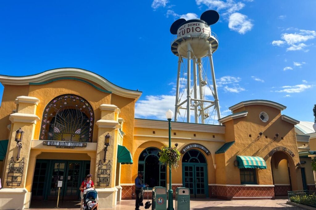 The image shows the entrance area of Walt Disney Studios Park at Disneyland Paris under a bright blue sky with scattered clouds. Dominating the background is the iconic white water tower topped with black Mickey Mouse ears and the words “Walt Disney Studios” written across the tank. In the foreground are warm yellow and cream-coloured art deco style buildings with curved rooflines, teal trim, and decorative windows. A few visitors walk through the open plaza area, including a person pushing a stroller and another standing near a rubbish bin and lamp post. Green awnings and hanging flower baskets add colour to the scene. The sunlight creates strong shadows across the pavement, while the bright colours and classic Hollywood-inspired architecture give the area a cheerful, cinematic feel associated with the Disney park entrance.