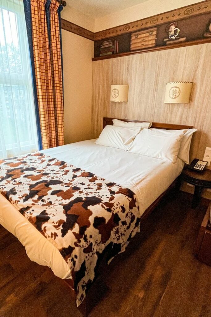 The image shows a cosy western-themed bedroom inside Disney Hotel Cheyenne at Disneyland Paris. A large double bed with crisp white bedding sits in the centre of the room, topped with a cow-print blanket in shades of brown, black, and cream that reinforces the cowboy-inspired design. The room features warm beige walls, dark wooden furniture, and wood-style flooring that give the space a rustic feel. Above the bed are two matching wall lamps decorated with sheriff badge designs, while a decorative border near the ceiling includes western-style patterns and small character artwork. A large window with yellow and blue checked curtains allows natural light to brighten the room. A bedside table with a telephone sits beside the bed, and the overall atmosphere feels comfortable, simple, and themed to resemble a classic frontier lodge.