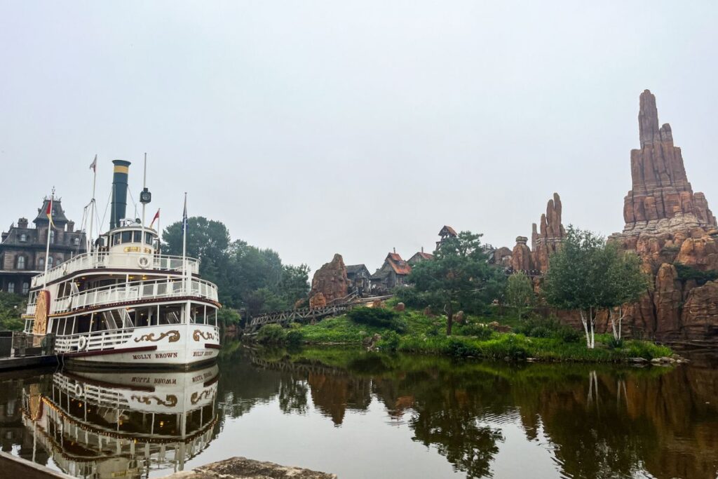 Frontierland The image shows a peaceful waterfront scene inside a western-themed area of a theme park on an overcast day. In the foreground, a large white paddle steamer named Molly Brown is docked along the edge of a calm river, with its multi-level decks, tall smokestack, and decorative gold trim reflected clearly in the still water below. Across the river, rugged orange and brown rock formations rise dramatically into the sky, creating the appearance of an Old West desert landscape. Wooden tracks and small rustic buildings are partially visible among the rocks and greenery, suggesting a nearby roller coaster attraction. Trees, shrubs, and patches of grass line the riverbank, softening the rocky scenery. The grey sky and glassy reflections on the water give the image a quiet, atmospheric feel despite the large-scale theme park setting.