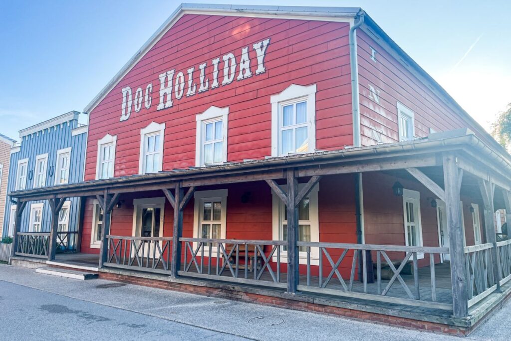 The image shows a large western-style hotel building at Disney Hotel Cheyenne in Disneyland Paris on a bright, sunny day. The two-storey structure is painted deep red with white trim around the windows and roofline, designed to resemble an old frontier town building from the American Wild West. Across the upper front of the building, large white lettering spells out “Doc Holliday,” referencing the famous cowboy and gunslinger. A long wooden porch with rustic railings wraps around the front and side of the building, supported by dark wooden beams that enhance the frontier theme. Several white-framed windows line both floors, while neighbouring pastel blue hotel buildings are partially visible beside it. The sky above is clear and bright blue, with sunlight creating soft reflections on the building and casting gentle shadows across the quiet pathway in front of the hotel.