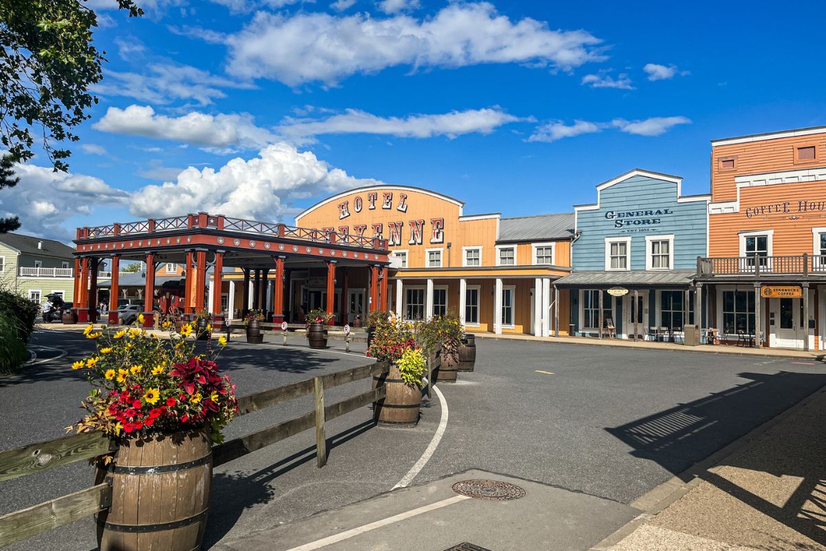 The image shows the exterior entrance area of Disney Hotel Cheyenne at Disneyland Paris on a bright sunny day. The hotel is designed to resemble an Old West frontier town, with wooden-style buildings painted in warm beige, cream, and muted pastel colours. In the centre stands a large covered entrance with dark red wooden beams and a sign reading “Hotel Cheyenne” above the doorway. Decorative wooden barrels filled with colourful flowers line the curved roadway and pathways leading toward the entrance. The sky is vivid blue with scattered white clouds, creating a bright and welcoming atmosphere. The surrounding area is clean and spacious, with wide pavements, wooden fencing, and western-themed architectural details throughout. The overall image captures the rustic cowboy-inspired style of the hotel while still feeling family-friendly, cheerful, and well maintained.