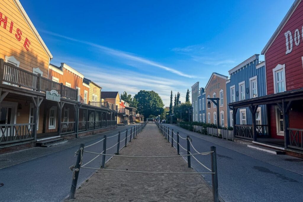 The image shows a quiet street scene inside Disney Hotel Cheyenne at Disneyland Paris, designed to resemble an old western frontier town. Colourful wooden buildings line both sides of the street in shades of red, blue, yellow, and orange, each featuring balconies, porches, and rustic signage inspired by saloons and frontier stores. Rope barriers run along the centre of the wide pathway, creating the appearance of an old dirt road through a Wild West town. The architecture includes wooden railings, covered walkways, and decorative details that reinforce the cowboy theme throughout the hotel area. The sky above is bright blue with soft evening light casting long shadows along the street. With very few people visible, the scene feels peaceful and atmospheric, capturing the immersive western styling and relaxed environment of the themed Disney hotel.