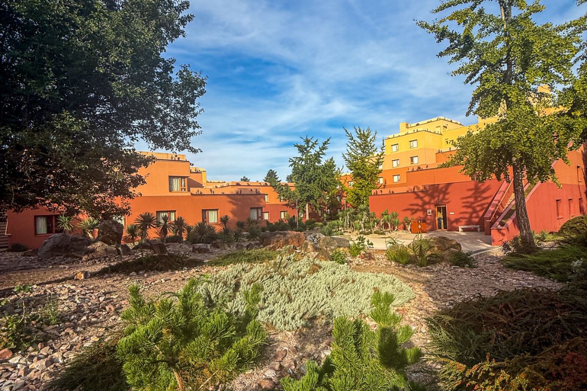 The image shows the exterior courtyard area of Disney Hotel Santa Fe at Disneyland Paris on a warm, sunny day. Several low-rise hotel buildings painted in shades of terracotta, burnt orange and sandy yellow surround a landscaped desert-style garden inspired by the American Southwest. The foreground is filled with pale gravel, large natural rocks, succulents and drought-tolerant plants arranged in winding patterns that resemble a desert landscape. Tall green trees frame the scene on both sides, adding shade and contrast against the warm-coloured buildings. Small pathways weave through the courtyard, leading toward hotel entrances and seating areas. The sky above is bright blue with thin wisps of cloud, while soft afternoon sunlight casts long shadows across the rocks and pathways, creating a calm and peaceful atmosphere around the hotel grounds.
