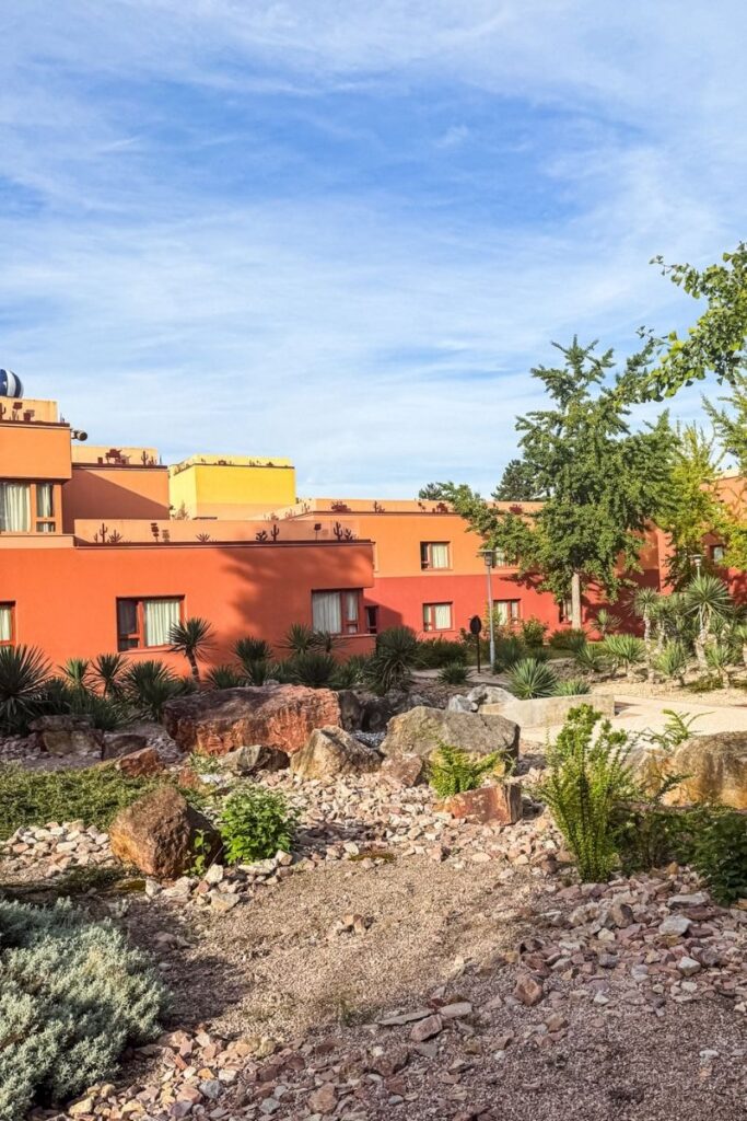 Hotel Santa Fe Exterior The image shows a landscaped outdoor area at Disney Hotel Santa Fe in Disneyland Paris, designed to resemble the desert scenery of the American Southwest. In the foreground, pale gravel and rocky pathways are scattered with large reddish-brown boulders, small shrubs and desert-style plants including yucca and succulents. Beyond the garden, low-rise hotel buildings painted in warm shades of orange, terracotta and pale yellow stretch across the background. The flat rooftops feature decorative silhouettes of cacti and desert motifs along the edges, reinforcing the Route 66 and Cars-inspired theme of the hotel. Tall trees and shrubs soften the edges of the buildings while bright sunlight illuminates the scene. A pale blue sky with thin clouds hangs overhead, creating a warm, dry atmosphere that resembles a desert resort despite being located in Disneyland Paris.