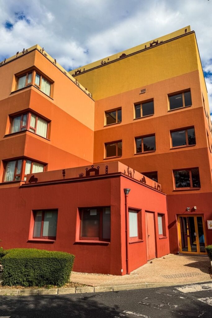 Hotel Santa Fe Exterior The image shows one of the exterior hotel buildings at Disney Hotel Santa Fe in Disneyland Paris. The multi-storey structure is painted in warm desert-inspired colours, blending shades of terracotta orange, deep rust red and sandy yellow. Rows of square windows with dark frames line the building, while decorative silhouettes of cacti, road signs and desert motifs run along the roofline, reflecting the Route 66 and Disney Pixar Cars theme of the hotel. The entrance area is visible at ground level with glass doors and a small paved pathway leading inside. A neatly trimmed hedge sits beside the building, while sunlight casts warm highlights and shadows across the textured walls. Above, large white clouds drift across a blue sky, giving the scene a bright and welcoming feel that matches the relaxed desert motel aesthetic throughout the hotel.