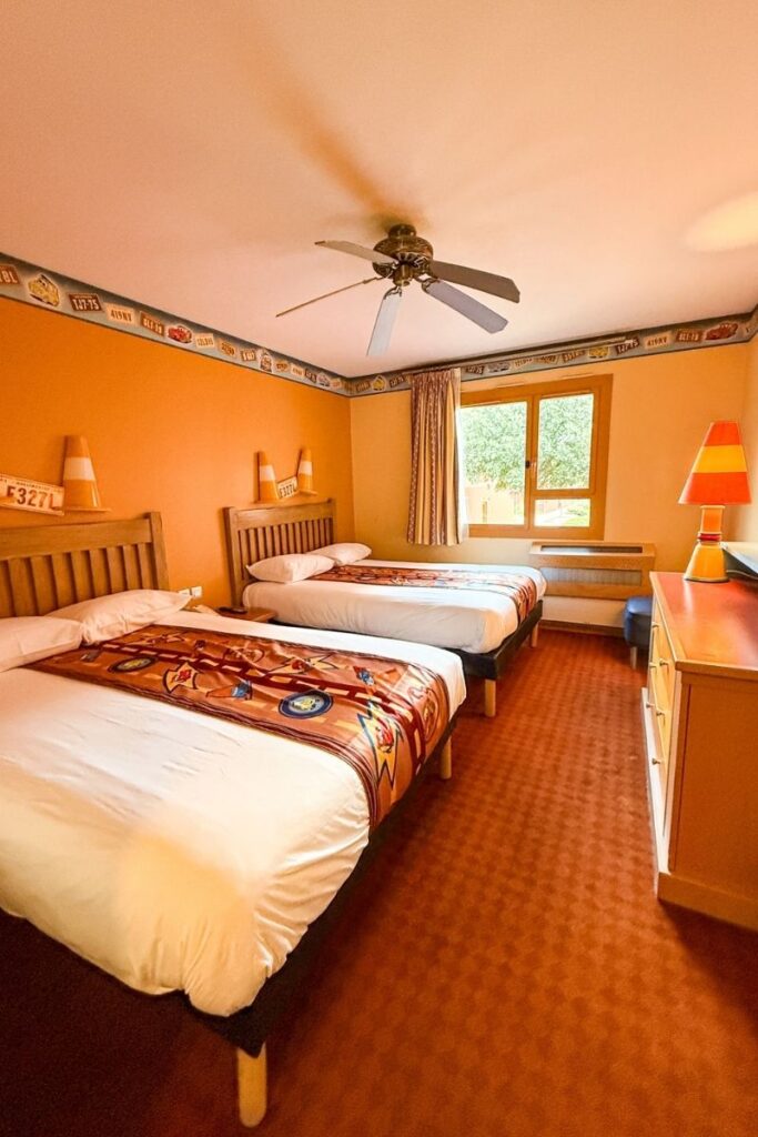 Hotel Santa Fe Room The image shows a family hotel room at Disney Hotel Santa Fe decorated with a Disney Pixar Cars-inspired theme. Two double beds with wooden headboards stand side by side against warm orange walls, each topped with white bedding and colourful runners featuring Cars characters and Route 66 designs. Matching traffic-cone-shaped lamps sit on shelves above the beds, adding playful themed details to the room. A ceiling fan hangs centrally above, while patterned wallpaper borders the upper walls with road signs and motel-inspired graphics. On the right side of the room, a large window lets in natural daylight through striped curtains, brightening the cosy interior. A dresser with another themed lamp sits beneath the window, and the carpet is a warm rust-red colour that complements the desert-inspired palette used throughout the hotel room.