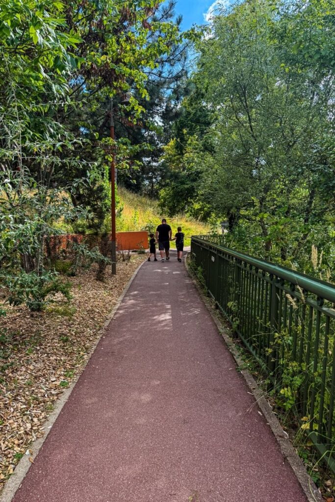Santa Fe to Disneyland The image shows a quiet pedestrian pathway surrounded by dense greenery and mature trees on a bright summer day. A smooth reddish walking path stretches through the centre of the scene, gently curving into the distance as it disappears into the landscaped surroundings. On the right side, a dark green metal railing runs alongside the path, partially covered by tall grasses and leafy plants. To the left, woodchip-covered ground sits beneath shrubs and trees with branches extending overhead, creating patches of shade along the walkway. In the distance, an adult walks hand in hand with two children, adding a sense of scale and calm family atmosphere to the image. Sunlight filters through the trees, illuminating the foliage in shades of green while a soft blue sky with scattered clouds is visible through the canopy above.