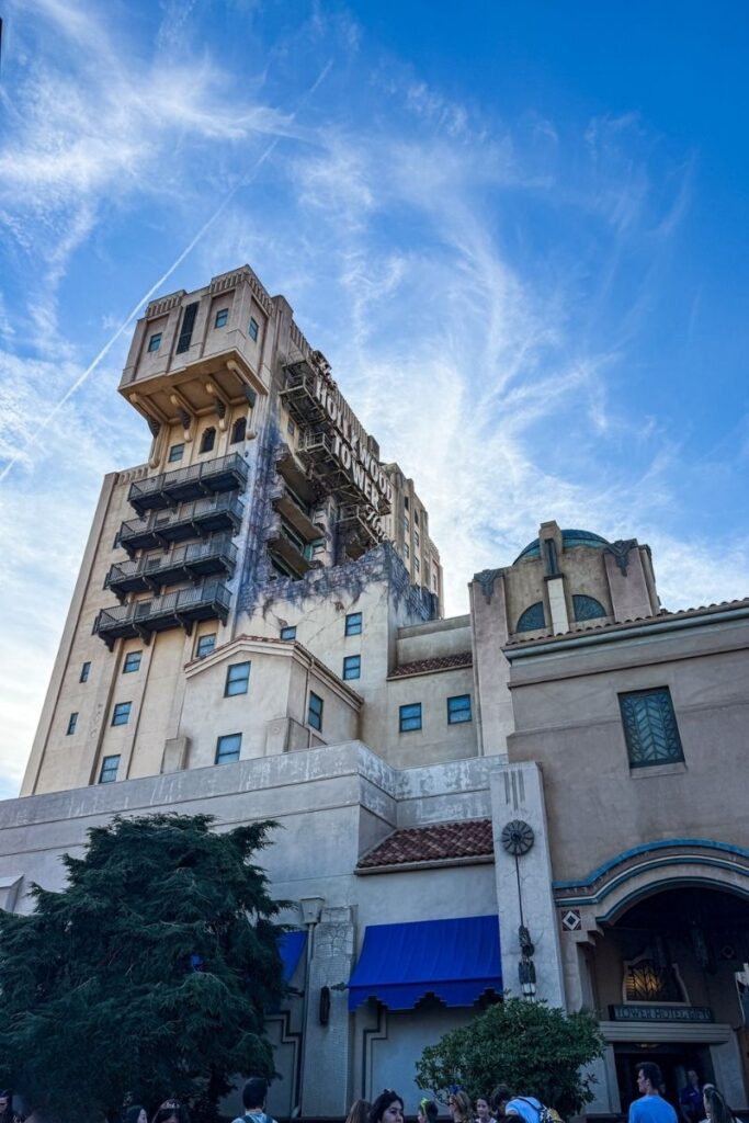 Tower of Terror The image shows a tall theme park hotel attraction designed in an aged Art Deco style, viewed from ground level against a bright blue sky streaked with thin white clouds. The beige-coloured tower rises high above the surrounding buildings and features balconies, narrow windows, and weathered detailing that gives it a slightly abandoned appearance. Large faded lettering reading “Hollywood Tower Hotel” is visible near the top of the structure. In the foreground, smaller connected buildings include arched entrances, textured walls, and blue awnings above doorways and windows. Trees and shrubs partially frame the lower part of the image, while visitors gather near the entrance at ground level. The upward perspective emphasizes the height and imposing nature of the building, creating a dramatic and slightly eerie atmosphere.