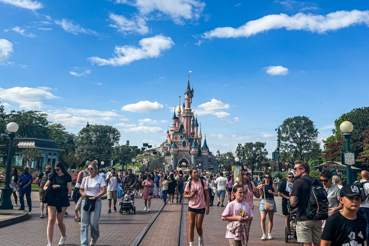 The image shows a wide view down the centre of Main Street at Disneyland Paris on a bright, sunny day. In the distance, the pink Sleeping Beauty Castle stands prominently at the end of the street, framed by trees and decorative lampposts on both sides. The sky above is vivid blue with scattered white clouds, creating a bright and cheerful atmosphere. Tram tracks run through the middle of the brick-paved street, leading the eye toward the castle. Large crowds of visitors fill the scene, including families with pushchairs, groups of friends, and children walking around the park. Some people are dressed casually in shorts and T-shirts, while others wear Disney-themed clothing and accessories. The overall image captures the lively energy and busy atmosphere of a popular day inside Disneyland Paris.