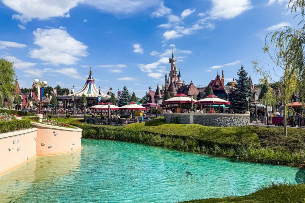 The image shows a picturesque section of Disneyland Paris on a bright, sunny day, viewed across a clear turquoise lagoon in the foreground. The calm water reflects the blue sky and scattered white clouds above, while neatly trimmed grass and landscaped banks frame the edge of the lagoon. Beyond the water, colourful red-and-white parasols shade outdoor seating areas where guests gather and relax. To the left, a whimsical carousel-style attraction with a striped canopy adds vibrant colour and movement to the scene. In the background, the pink spires and ornate turrets of Sleeping Beauty Castle rise above the surrounding buildings, creating a fairytale focal point. Decorative trees, flowerbeds and themed architecture fill the area, while visitors stroll through the park enjoying the attractions. The image captures the lively yet scenic atmosphere of Disneyland Paris on a clear summer day.