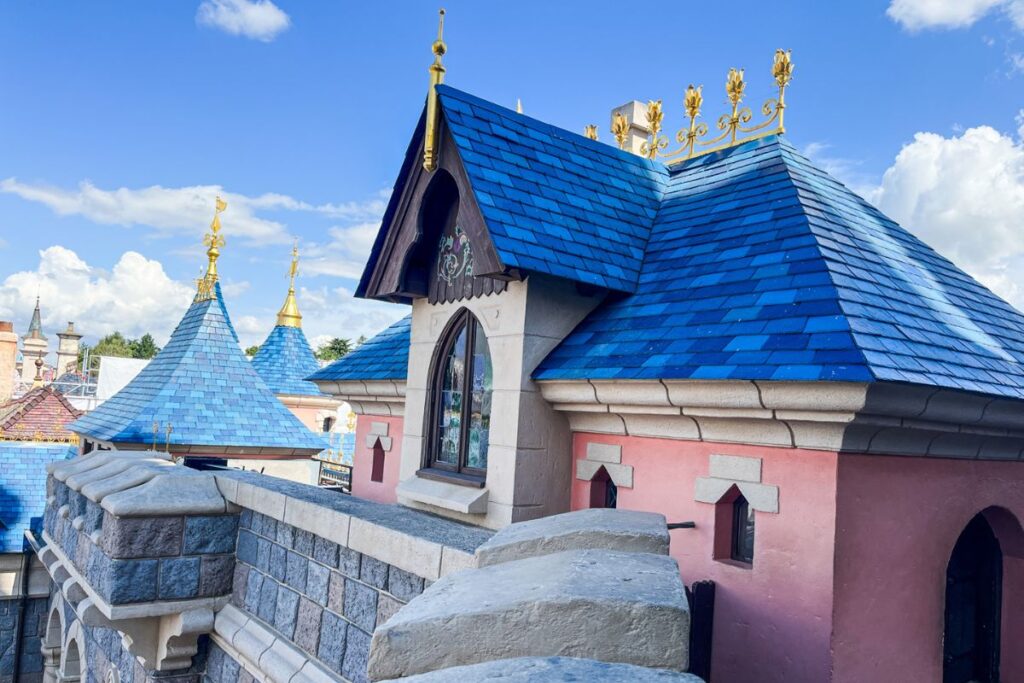 Disneyland Paris Castle Balcony A close-up view of the upper architecture of Sleeping Beauty Castle at Disneyland Paris, showing a pink and pale stone turret with a striking bright blue tiled roof. Ornate gold decorative details line the roof ridges and several pointed turrets, which rise against a vivid blue sky filled with fluffy white clouds. In the foreground, a grey stone castle wall with a wide crenellated parapet runs across the image. Additional blue-roofed turrets and parts of the castle are visible behind it. The architecture combines fairytale-inspired shapes, colourful roofs, carved stonework and gold details. The image highlights the intricate decorative features and elevated walkways of Disneyland Paris’s iconic castle.