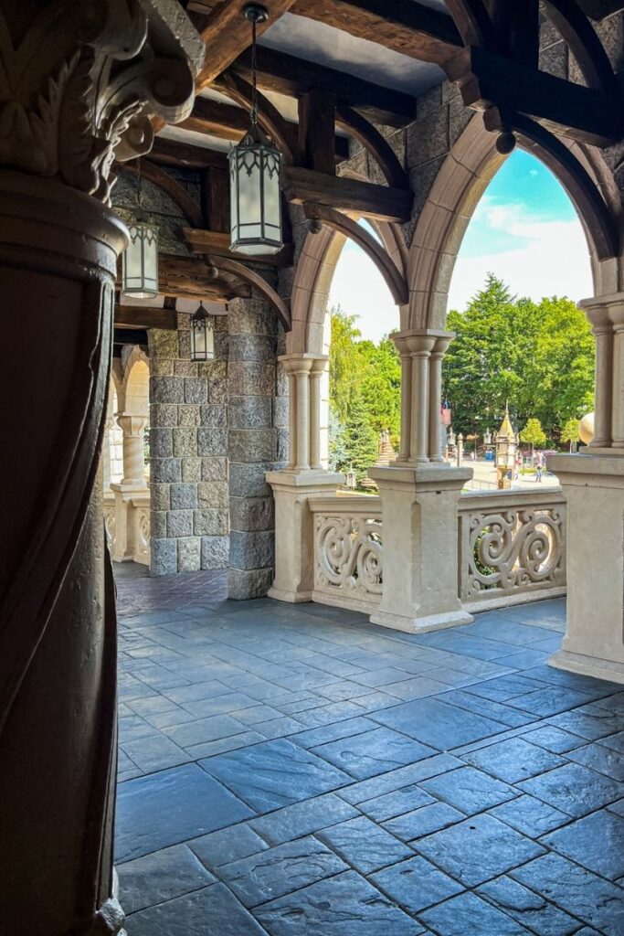 Disneyland Paris Castle Hallway An ornate covered walkway inside Sleeping Beauty Castle at Disneyland Paris, featuring grey stone walls, pointed Gothic-style arches and a series of decorative hanging lanterns. Thick dark wooden beams cross the ceiling, contrasting with the pale stonework. On the right, tall arched openings provide views over Disneyland Park, with trees and colourful park buildings visible outside in bright sunshine. A decorative stone balustrade runs beneath the arches. The floor is paved with large blue-grey stone slabs, while carved columns and intricate architectural details add to the medieval fairytale atmosphere. The corridor is relatively quiet and open, showcasing the detailed castle architecture and providing a glimpse of the park beyond.