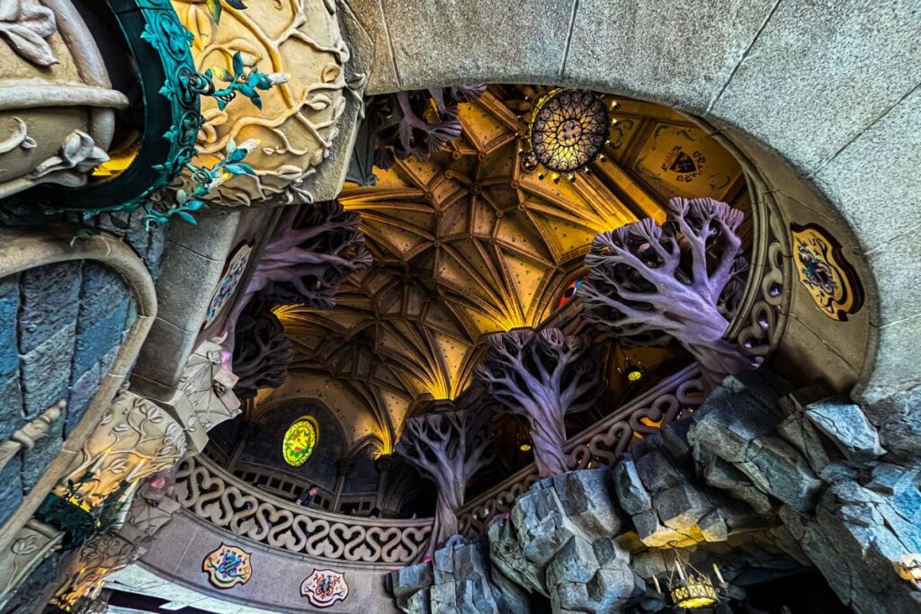 Disneyland Paris Castle Inside An interior view of Sleeping Beauty Castle at Disneyland Paris, looking upwards into an elaborate vaulted chamber. The ceiling features intricate wooden panels and decorative beams illuminated by warm golden lighting. Several tall, sculpted purple trees rise through the centre of the room, their branches spreading towards the ceiling and surrounding an upper balcony. Ornate stone railings with curved decorative patterns line the balcony, while grey rockwork forms the walls and lower sections of the scene. Hanging lanterns provide additional illumination. Decorative shields and colourful details are incorporated into the stonework. The unusual combination of medieval castle architecture, glowing woodwork and fantastical purple trees creates an immersive fairytale setting.