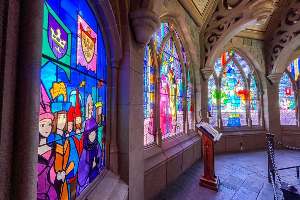 A section of Sleeping Beauty Castle at Disneyland Paris featuring a row of large, colourful stained-glass windows set within pale stone Gothic-style arches. The windows depict brightly coloured fairytale characters, royal figures, crowns and decorative heraldic symbols in shades of blue, pink, yellow, green and red. Sunlight shines through the glass, making the colours particularly vivid against the darker stone interior. A wooden information stand sits in front of the windows, while a low decorative barrier separates the viewing area from visitors. The room has a medieval-inspired design with carved stone arches and columns. The image highlights the detailed stained glass and fairytale storytelling incorporated into the castle’s interior.